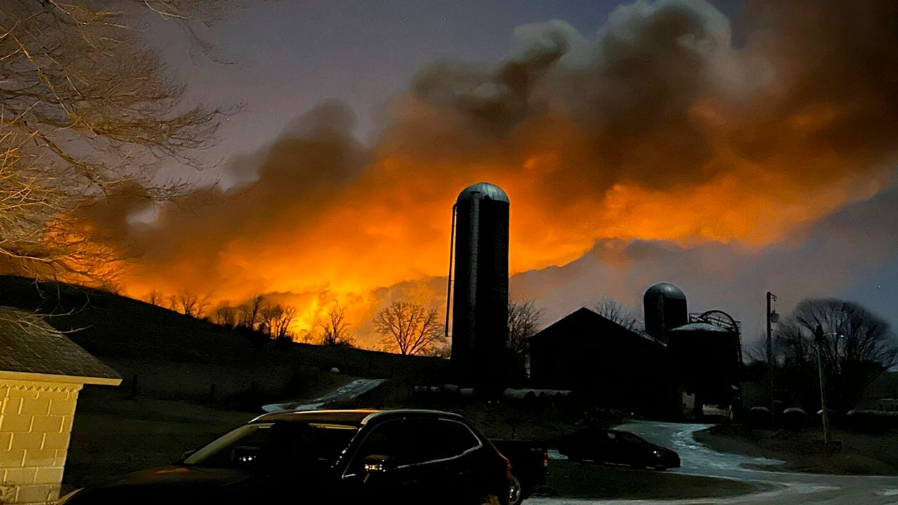  a large orange fire with black smoke rising in the air behind a farm and grain silo