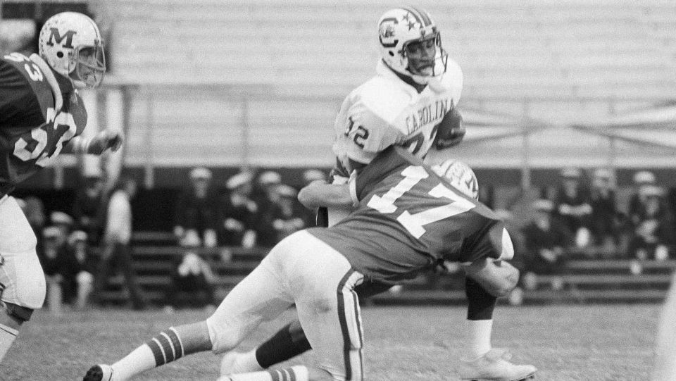 Three football players on the field in black and white. One attempting a tackle on another, with a third running behind. 