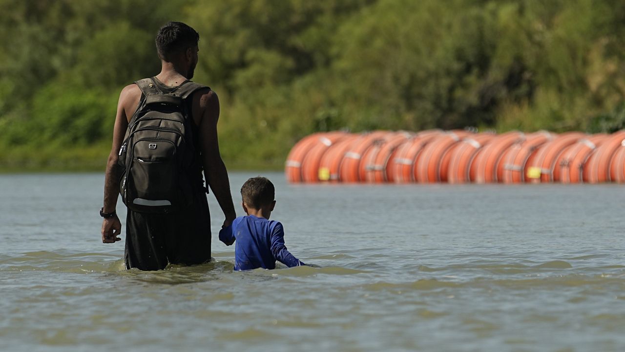 Migrants walk past large buoys being used as a floating border barrier on the Rio Grande, Aug. 1, 2023, in Eagle Pass, Texas. (AP Photo/Eric Gay)