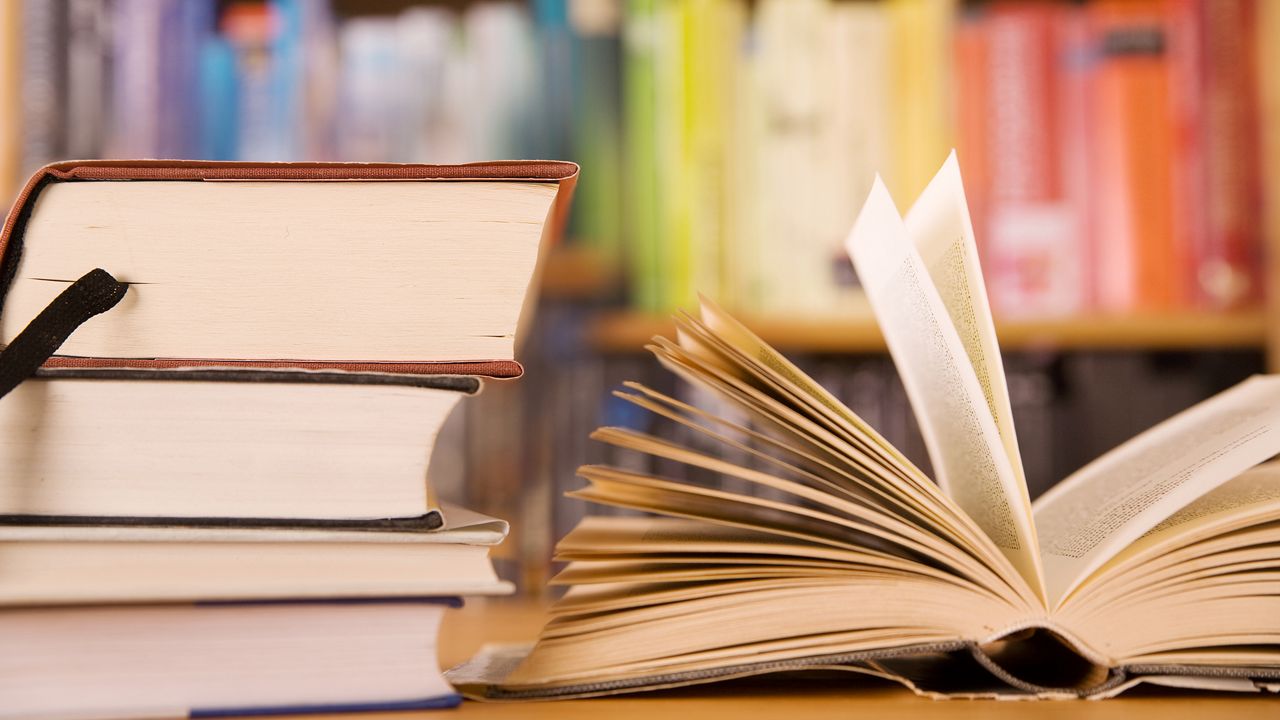 Stack of books and a open book on a wooden table. In the back rainbow sorted bookshelf. (Photo Credit: Getty Images)