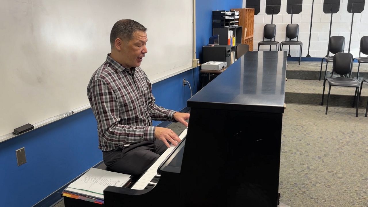 Bryan Long sits at a piano in the middle of the picture, he is looking down and singing while his fingers tap the keys.