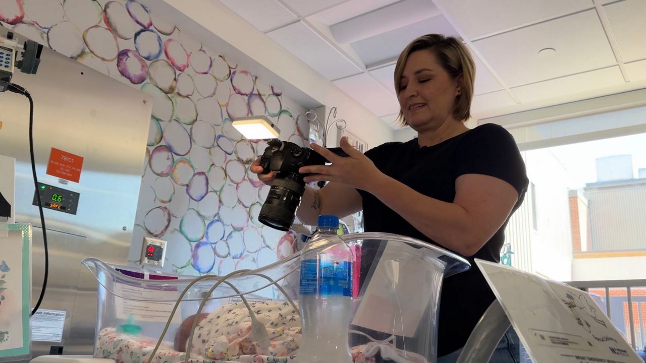 Mary Leach is taking a photo in the Novant Health NICU. She is smiling down at her camera, which is hovering over a small infant. Medical devices and cords are scattered throughout the space.