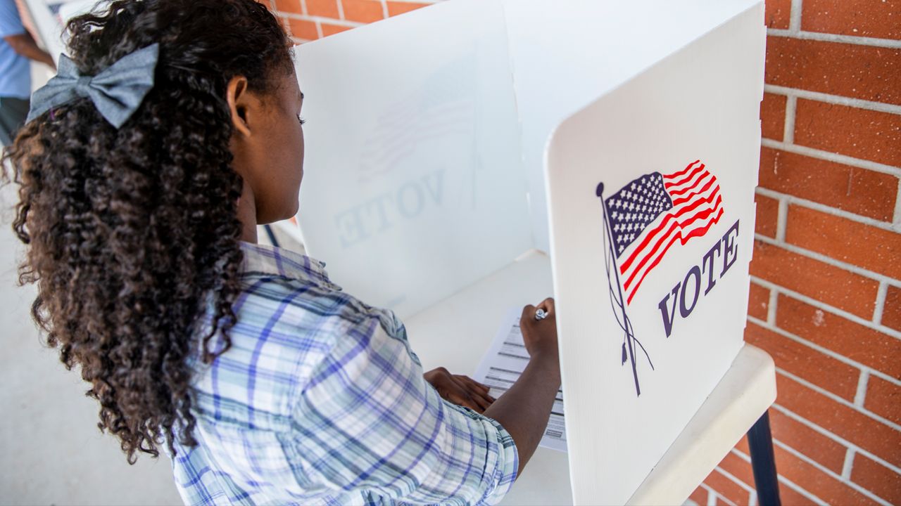 A woman filling out a ballot with partitions around her with the American flag and Vote painted on the side