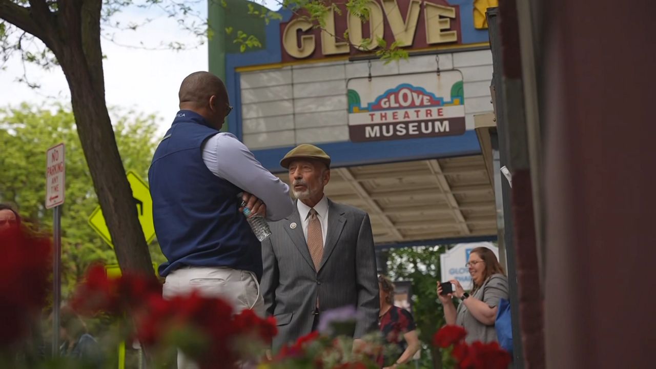 photo with flowers in the foreground, in background is a large sign hanging off of a building reading glove theatre museum, in front of hte sign are two men talking, next to them is a woman smiling and holding a phone poised to take a photo