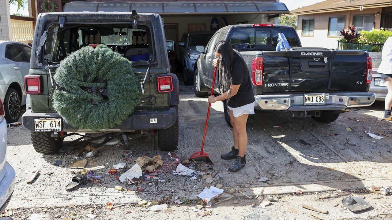 A woman sweeps debris from a driveway across the street from the home where a New Year's Eve fireworks explosion killed and injured people, Wednesday, Jan. 1, 2025, in Honolulu. (AP Photo/Marco Garcia)