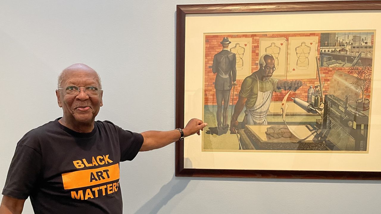 Robert Steele, an art collector, points at a lithograph by Ron Adams, titled “Blackburn,” that shows printmaker Robert Blackburn in his New York workshop where he mentored many printmakers. (Spectrum News/Michelle Broder Van Dyke)