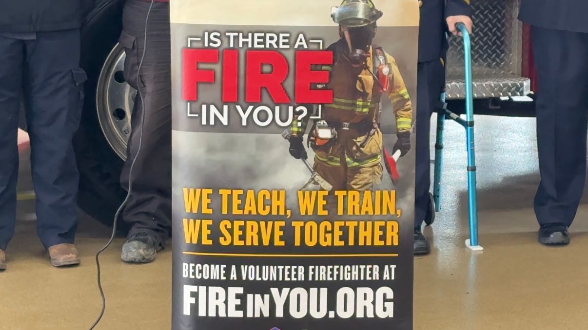 a volunteer firefighter recruitment sign in front of firefighters, who can only be seen by their legs and feet