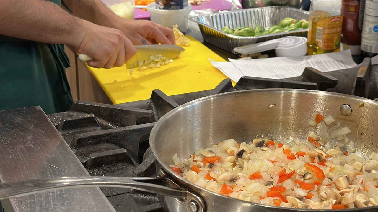 photo of a pan filled with food in the foreground and hands chopping food in the background 