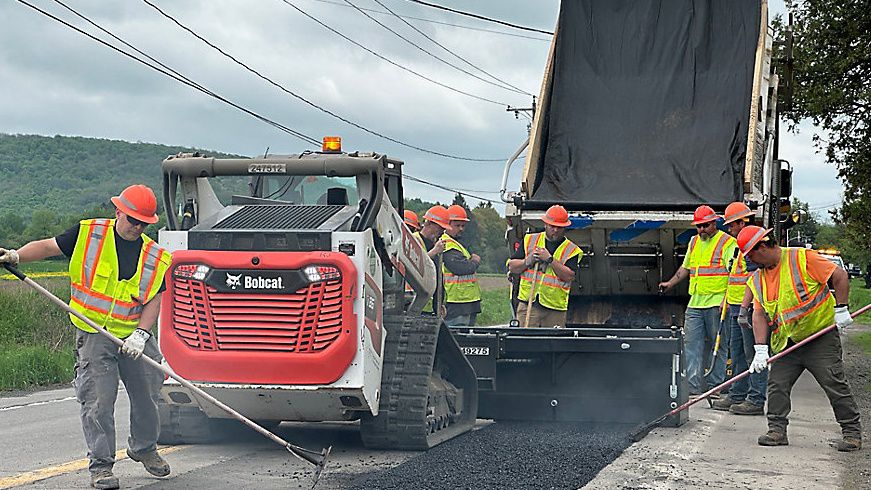 photo of a strip of fresh asphalt on the road, which is surrounded by 2 construction vehicles and several workers in hard hats and reflective vests using tools