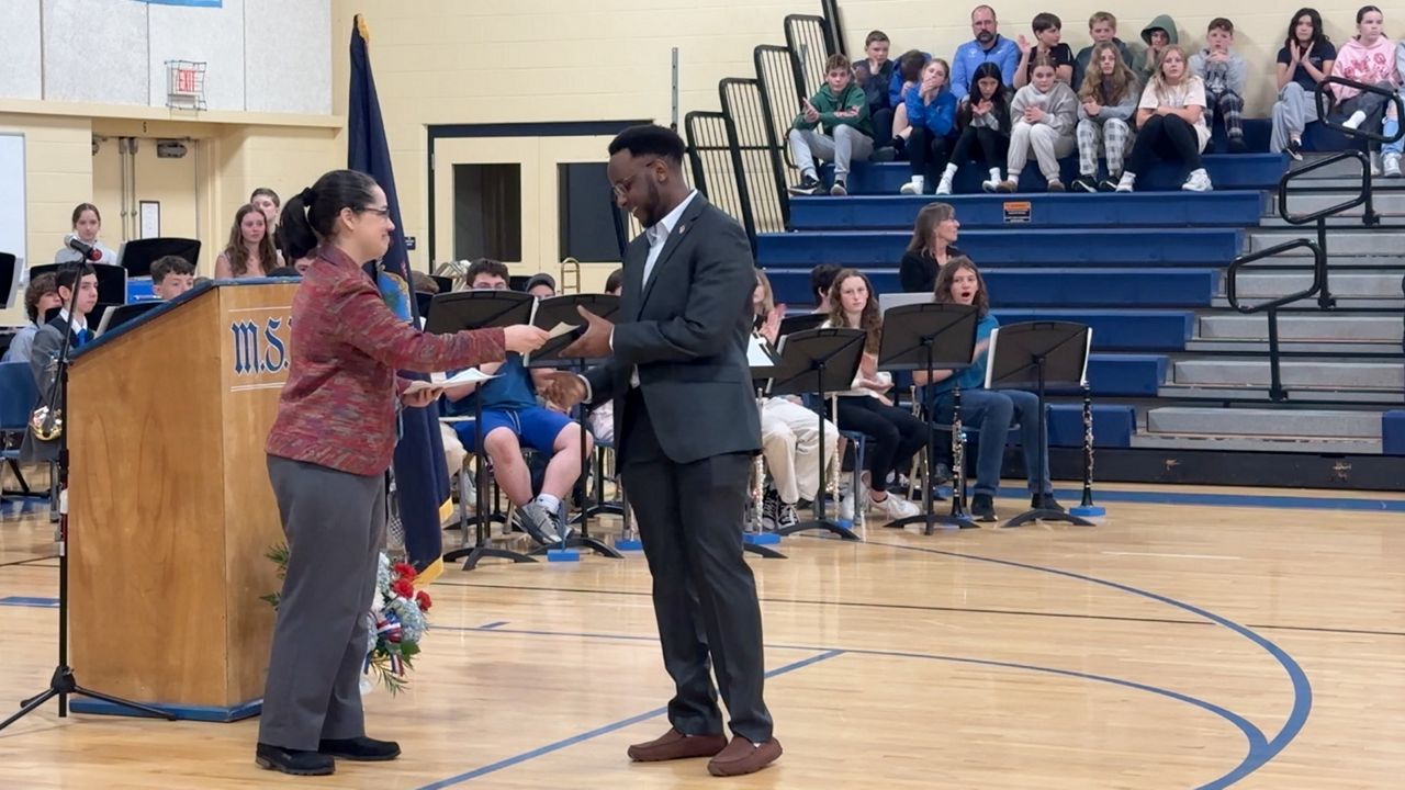 Ken Niyonkuru, 25, a native of Burundi now living in Biddeford, receives his certificate of US citizenship from Eriks Grunnet, acting Portland field office director of the US department of citizenship and immigration services, at a ceremony at Middle School of the Kennebunks on Friday, as students, faculty and staff watch. (Spectrum News/Sean Murphy)