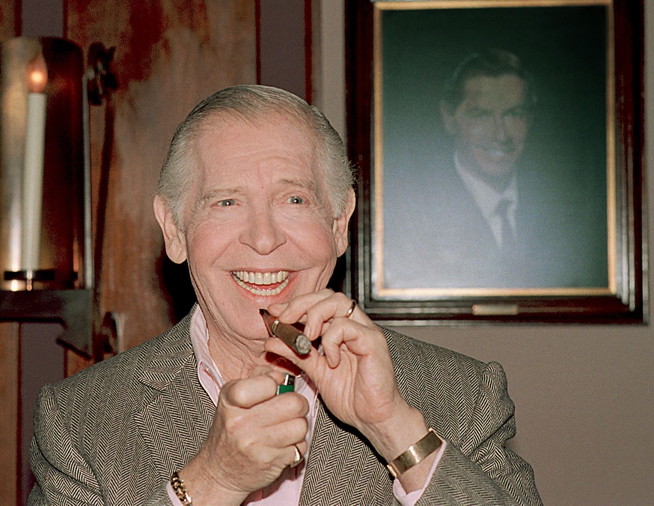 Milton Berle laughs as he lights a cigar during a luncheon at the Friars Club in Beverly Hills, Ca., Monday, July 11, 1988. Berle will celebrate his 80th birthday on July 12. At right is a portrait of Berle in his younger years. (AP Photo/Lennox Mclendon)