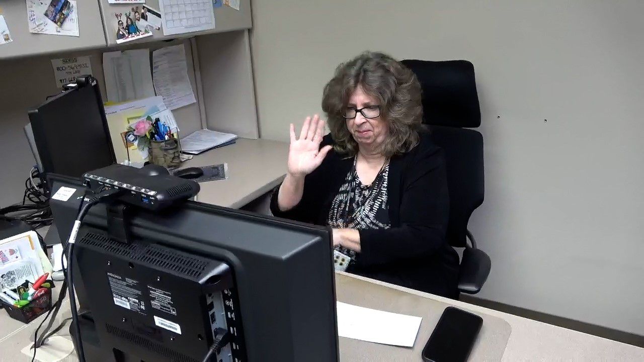 Beth Hostetler sits at her desk on a video phone call waving goodbye. 