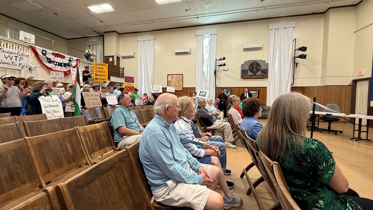 Protesters stand at the back of the room in Searsport’s municipal building as Susan Collins middle right prepares to deliver remarks. (Spectrum News/David Ledford)