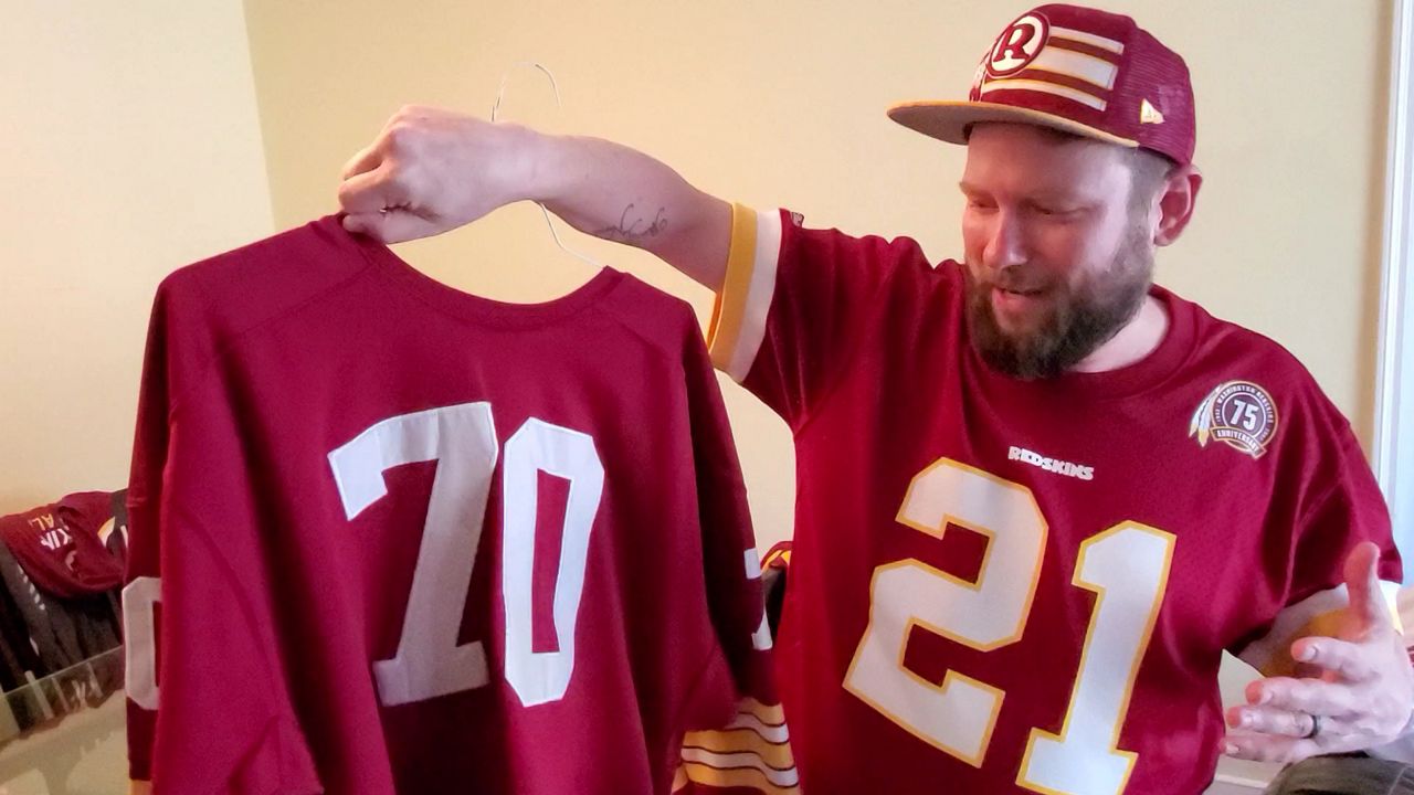 Bernie Linstrom holds up a Washington NFL team jersey while also wearing one and a team hat (Spectrum News)