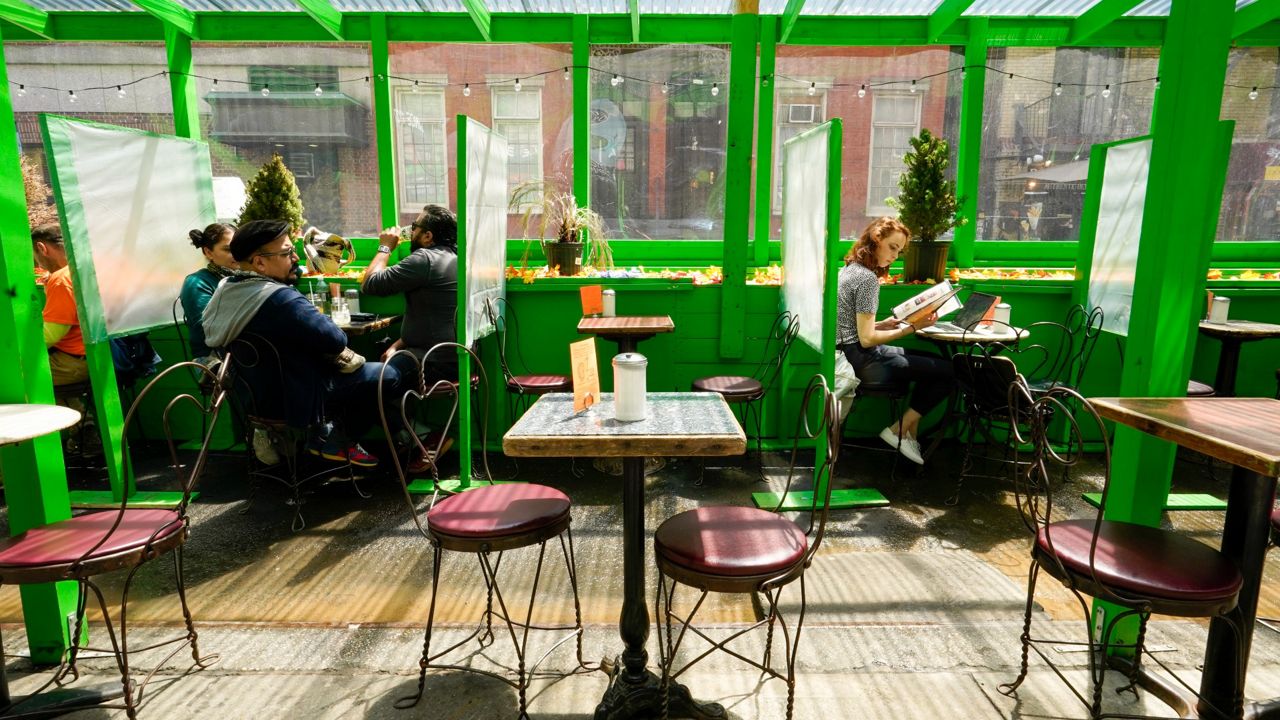 A wideshot of a green outdoor dining shed in front of NYC businesses and between parked cars.