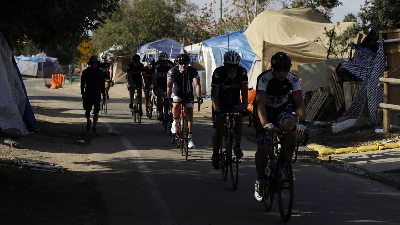 A group of cyclists pass the row of tents and tarps set up at a homeless encampment along the Santa Ana riverbed near Angel Stadium, Sept. 14, 2017, in Anaheim, Calif. (AP Photo/Jae C. Hong)