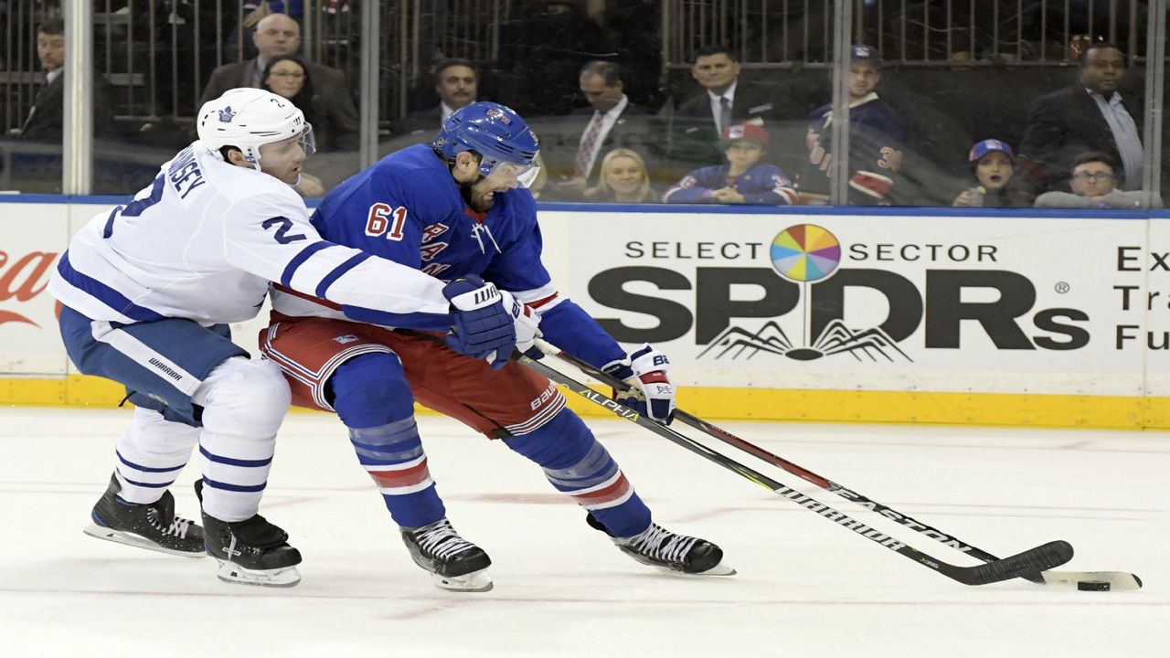 Ron Hainsey, left, in a white Maple Leafs uniform, swings his stick near a black hockey puck. Rick Nash, right, in a blue Rangers uniform, makes contact with the puck with the base of his stick.