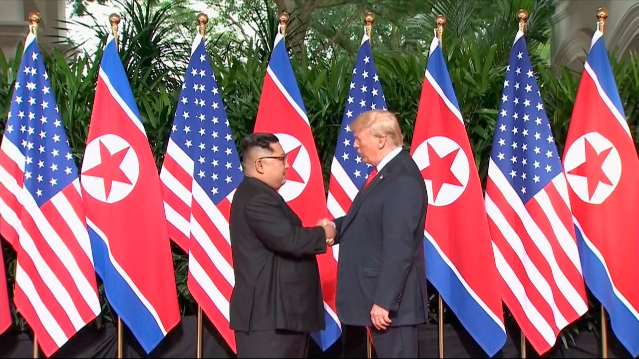 Kim Jong Un, left, wearing glasses and a black suit, shaking hands with Donald Trump, right, wearing a black suit, a white dress shirt, and a red tie. A row of American and North Korean flags are behind them.