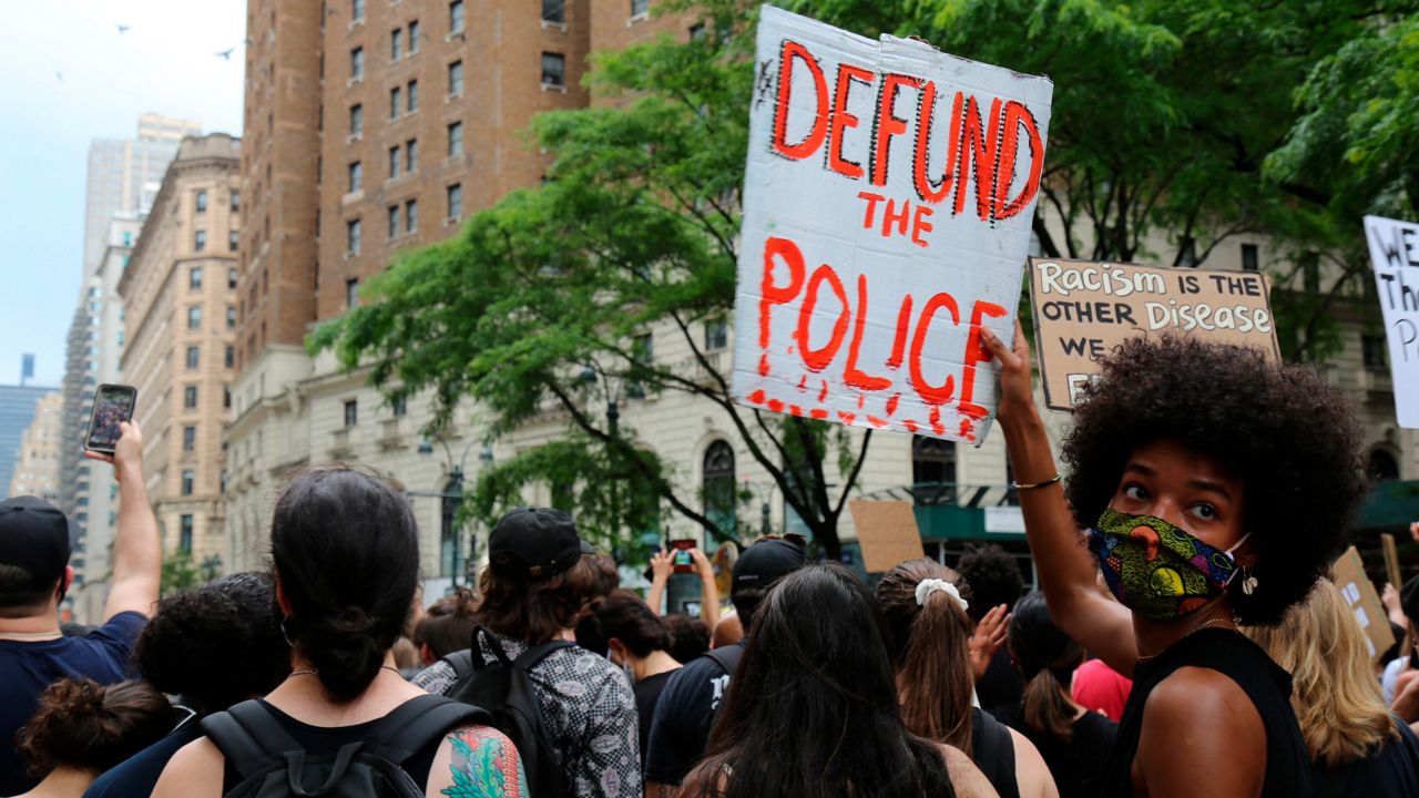 A woman holds a "defund the police" sign among a crowd of protesters as she looks over her shoulder.