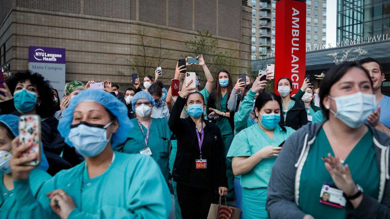 Medical personnel stand outside the NYU Langone Medical Center in medical uniforms with many people holding phones, documenting the 7 p.m. nightly applause celebrating essential workers.