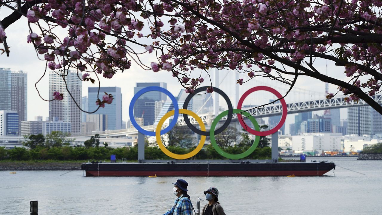 Under blooming cherry blossoms, people wearing protective masks to help curb the spread of the coronavirus walk with a backdrop of the Olympic rings floating in the water in the Odaiba section Thursday, April 8, 2021, in Tokyo. (AP Photo/Eugene Hoshiko)