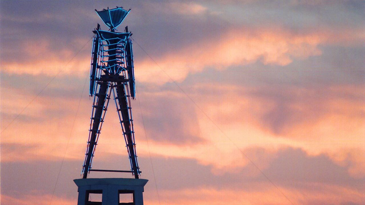 In this Aug. 26, 2002 file photo, the sun rises behind a wood and neon statue, the center piece of the annual Burning Man festival north of Gerlach, Nev. (AP Photo/Debra Reid, File)