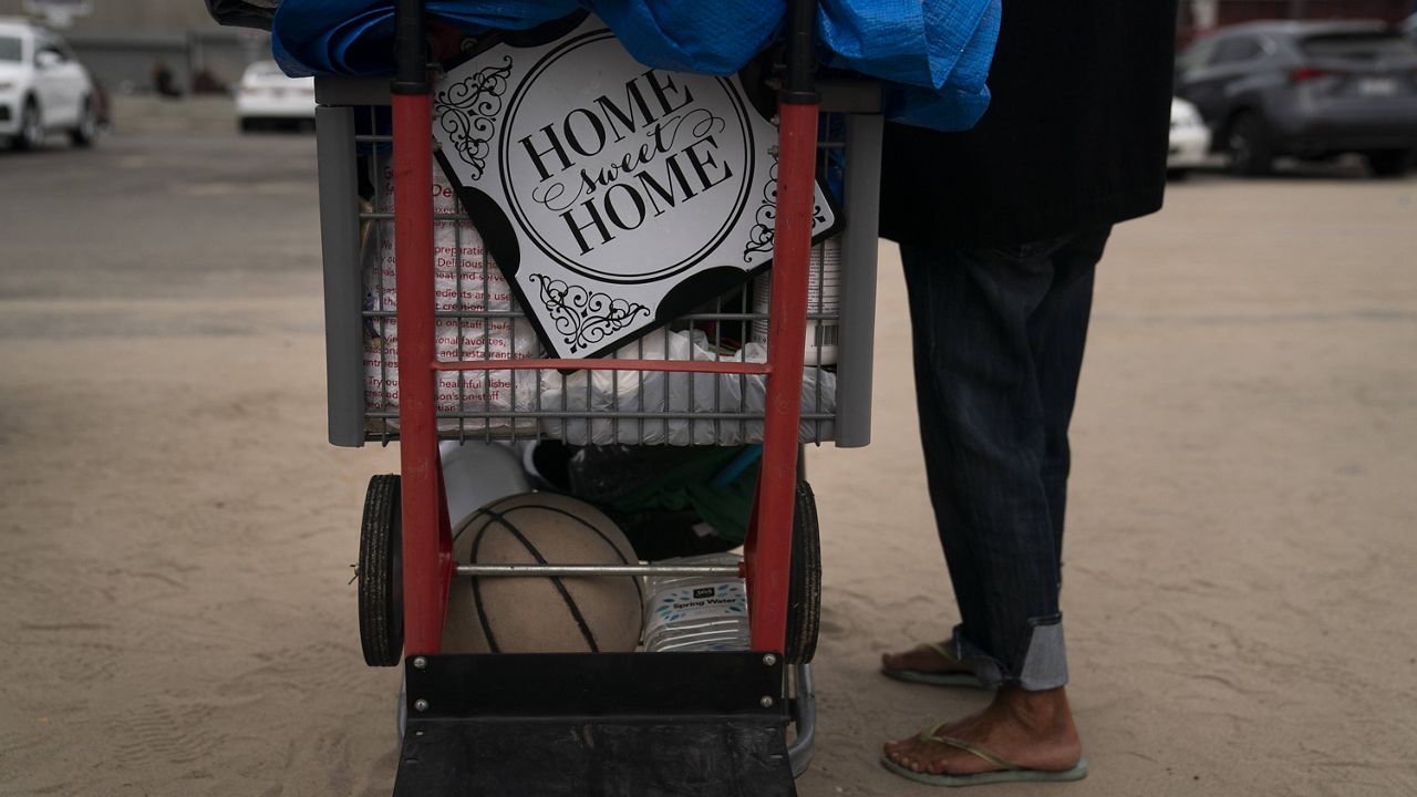 A homeless woman who goes by Ma Shanti stands next to her cart adorned with a sign that reads "Home Sweat Home" while waiting in line for a free meal in the Venice neighborhood of Los Angeles, Tuesday, June 29, 2021. (AP Photo/Jae C. Hong)