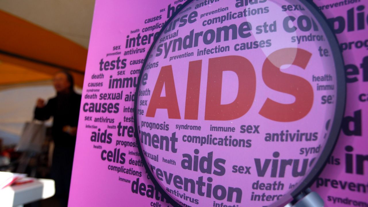 Close-up of a sign with AIDS in large red letters in center with a word cloud of related words surrounding, pink background and microscope over the center of the sign.