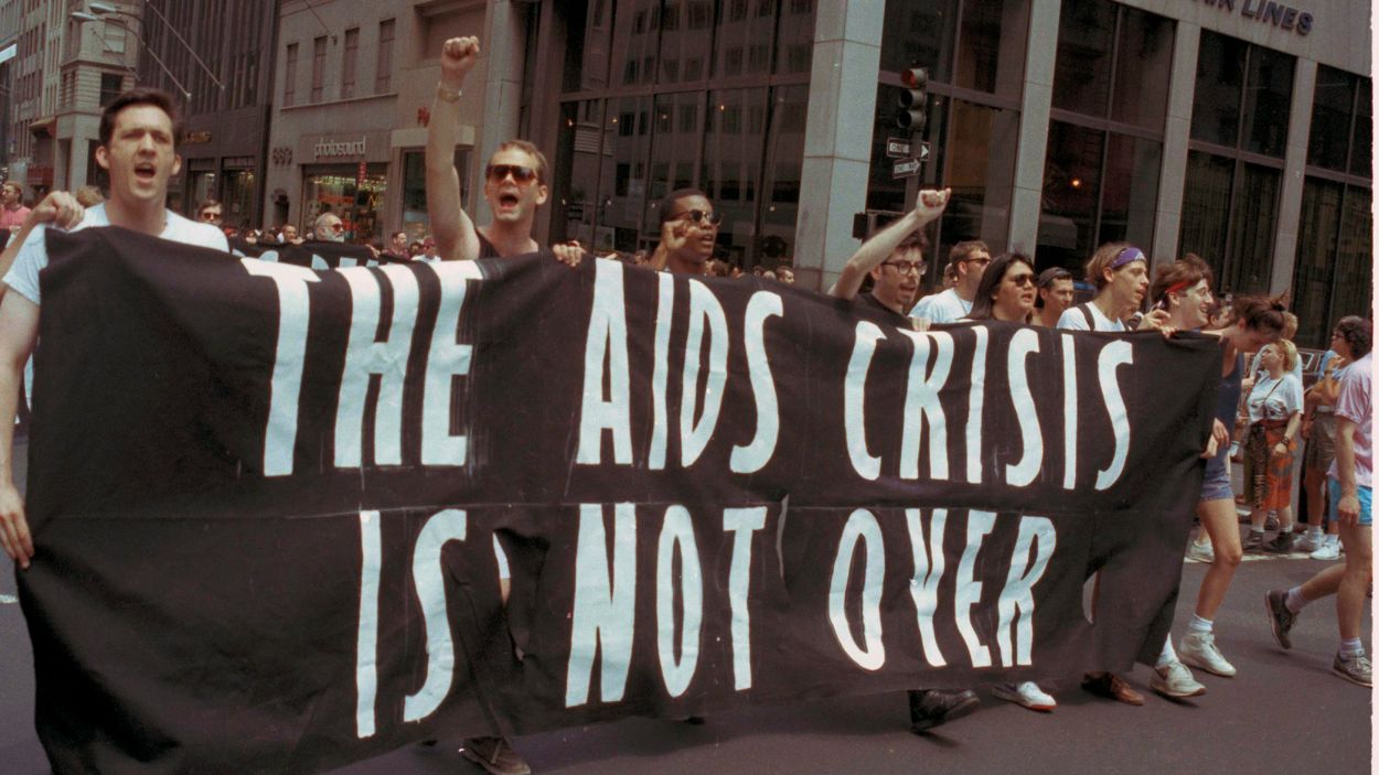 A group of marchers on the street during the 21st annual Gay Pride Parade in 1990 stand behind a black banner they're holding that reads in white letters: "The AIDS crisis is not over."