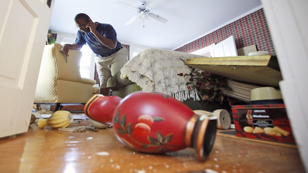 A red vase sits, overturned, on a hardwood floor. Broken glass and other vases are on the floor. A table is askew. A man leans against a chair while he holds a phone to his left ear.