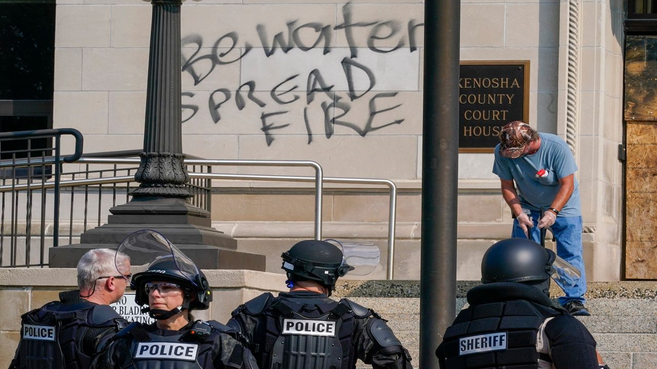 Volunteers sweep up debris as police in riot gear stand outside the Kenosha County Court House Monday, Aug. 24, 2020, in Kenosha, Wis. Protests broke out late Sunday night after a police shooting. (AP Photo/Morry Gash)