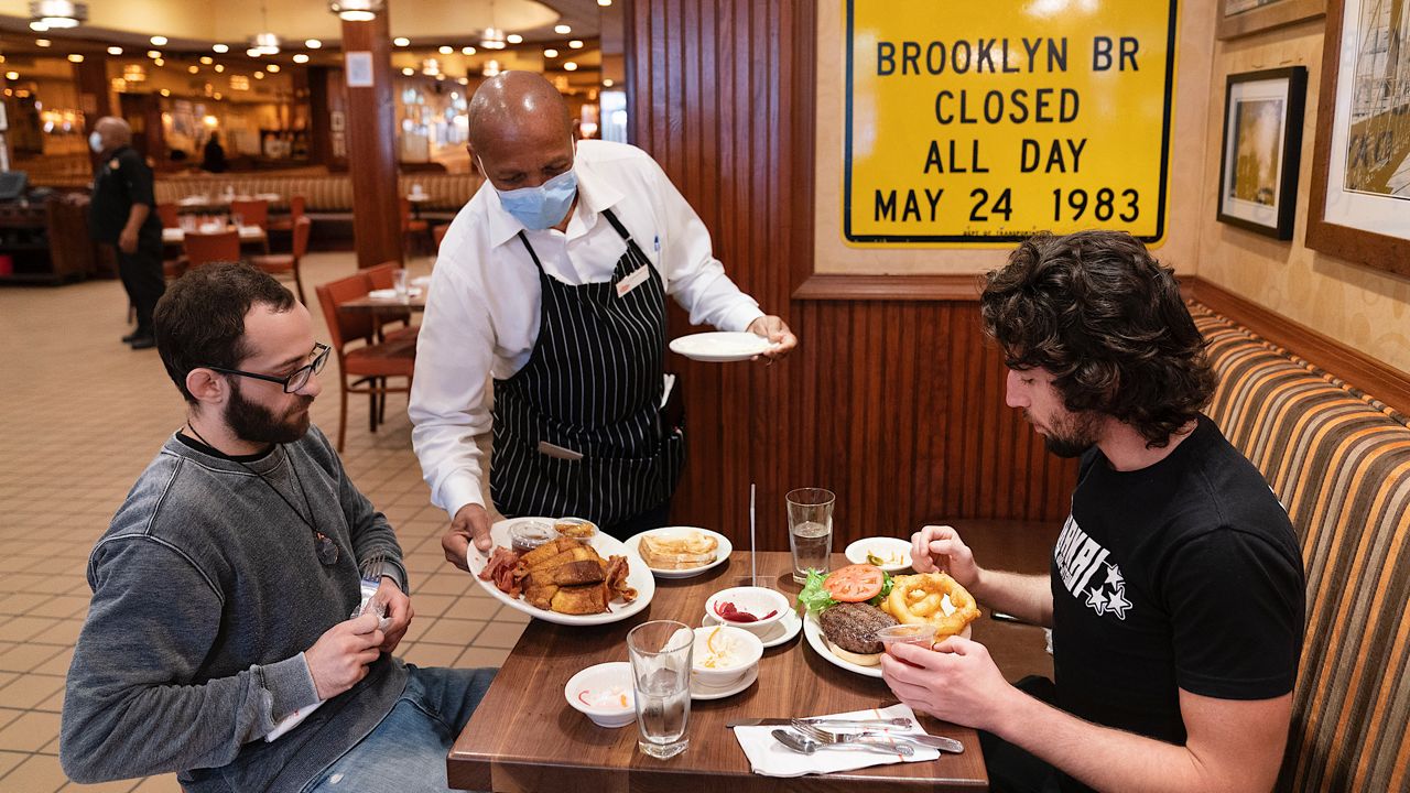 Waiter Lenworth Thompson serves lunch to David Zennario, left, and Alex Ecklin at Junior's Restaurant, Wednesday, Sept. 30, 2020 in New York. The city on Wednesday began allowing restaurants to offer indoor dining at 25 percent capacity. (AP Photo/Mark Lennihan)