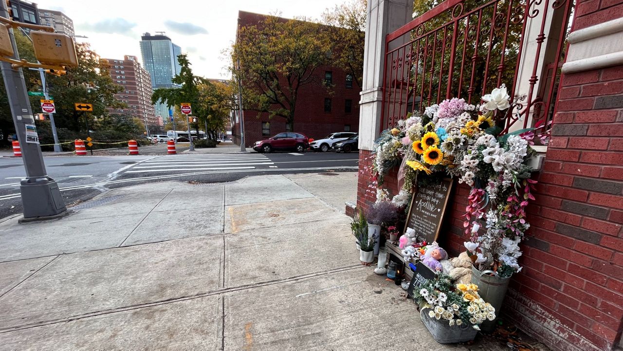 A memorial with flowers and a chalk board lays against a brick wall at the corner of an intersection, across the street from a block that is closed off to traffic. 