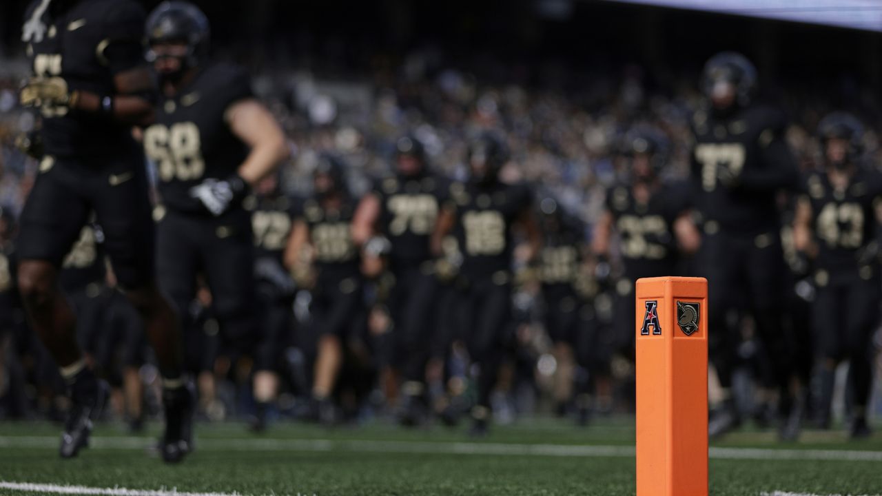 army football team runs onto field in background, blurry, with orange football pylon with army logos in focus in foreground