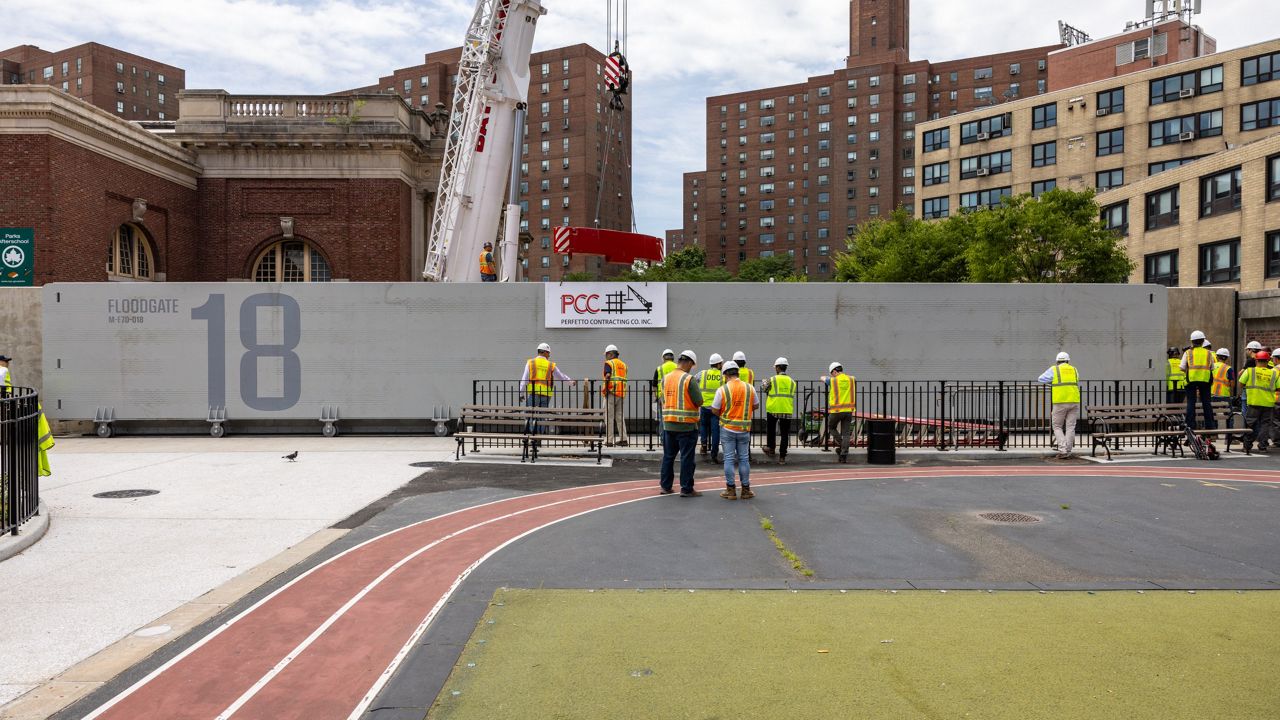 Construction crews standing in Asser Levy Park in Manhattan install a 10-foot-tall, 80-foot-long metal flood gate that slides across the middle of the park to prevent floodwaters from getting into the city.