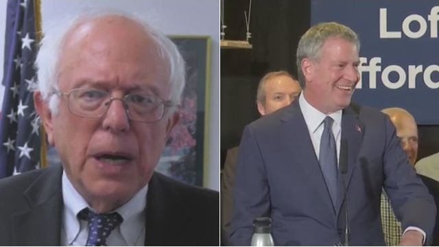 Vermont Sen. Bernie Sanders, left, and New York City Mayor Bill de Blasio, right. Sanders wears rectangular glasses, a black suit, and a purple tie. De Blasio wears a black suit, a white shirt, and a grey tie.