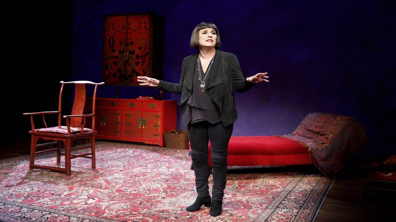 Eve Ensler, wearing a black fleece and a black shirt, and on a theater set with red furniture and a red carpet.