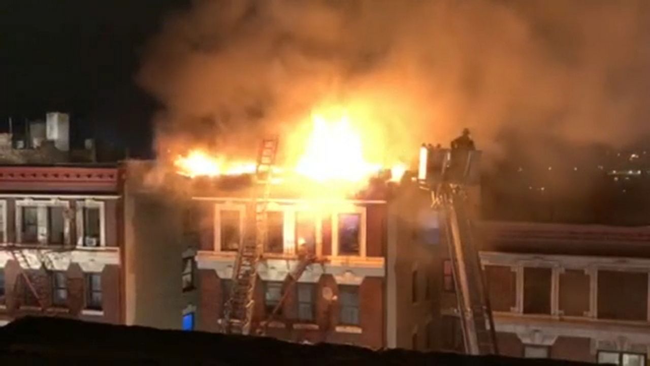 Flames are seen from the top of a building, sending smoke into the night sky. A firefighter ladder is near the blaze.