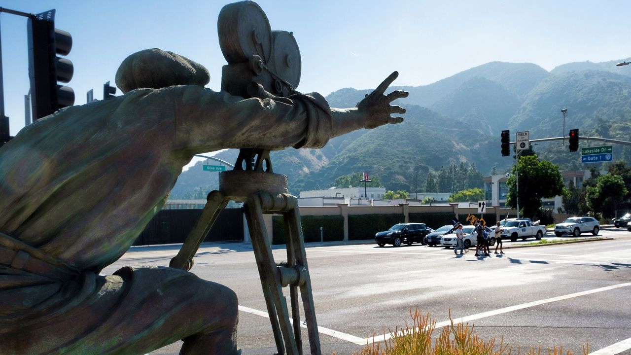 With a statue of a cameraman in the foreground, SAG-AFTRA picketers carrying signs cross the street are seen Tuesday near the gates of Warner Bros. studios in Burbank, Calif. (AP Photo/Richard Vogel)