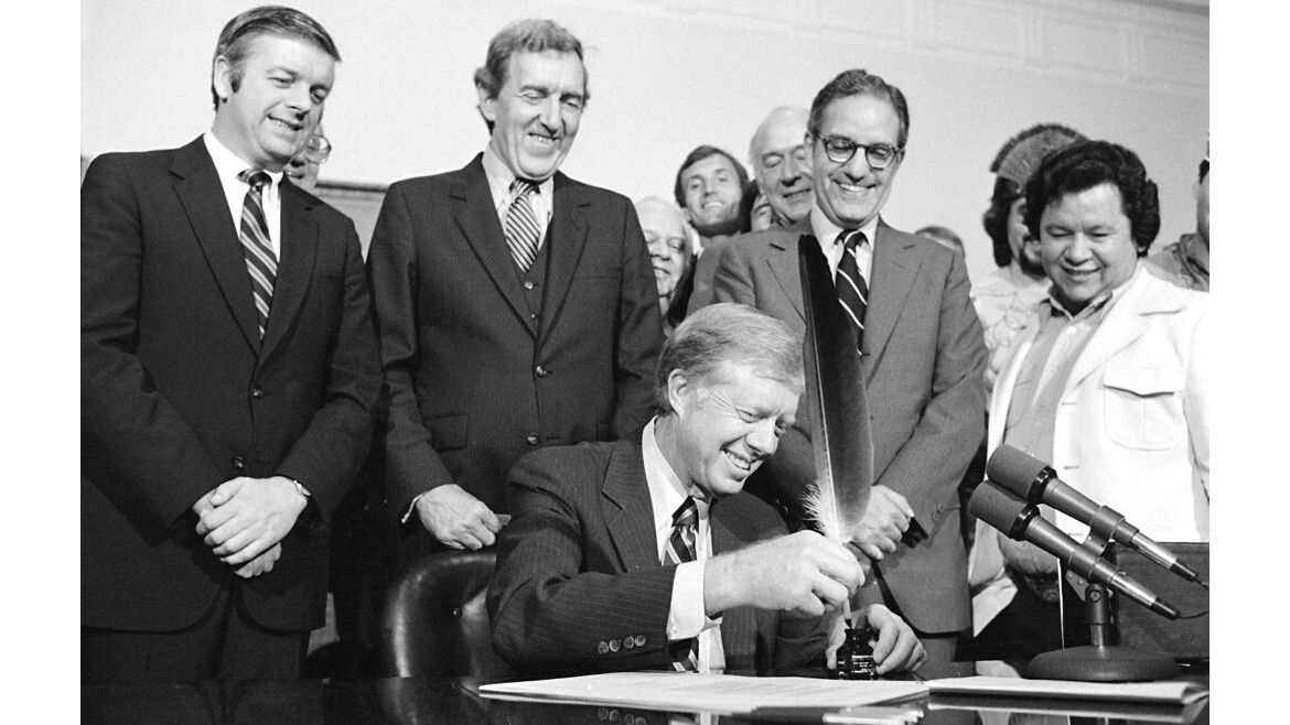 President Jimmy Carter uses an Indian quill pen to sign H.R.7919 the Maine Indian Claims Settlement Act of 1980 at a ceremony at the White House Friday, Oct. 10, 1980.  Shown in the photo are from left: Maine Governor Joseph Brennan, Secretary of State Edmund Muskie, Secretary of the Interior Cecil Andrus, Maine Senator George Mitchell, and Terrance Polchies.  (AP Photo/Barry Thumma)