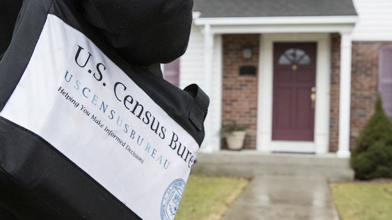 "U.S. Census Bur" in black print against a white paper on a blag back, held near a black jacket by someone standing near a front lawn and a red front door.