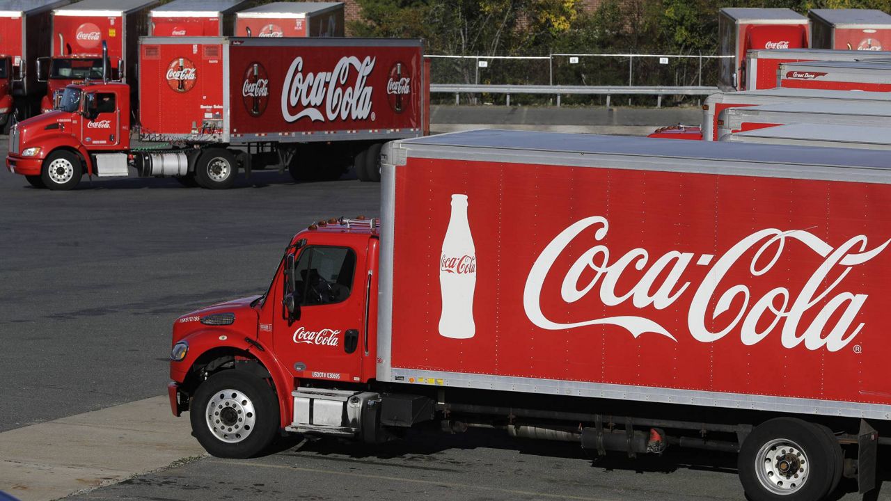 In this Oct. 14, 2019 photo a truck with the Coca-Cola logo, behind left, maneuvers in a parking lot at a bottling plant in Needham, Mass.  (AP Photo/Steven Senne)