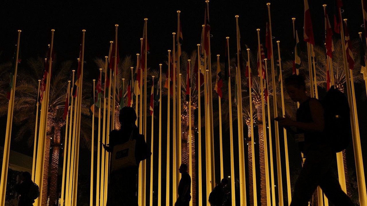 People walk near country flags during the COP28 U.N. Climate Summit, Friday, Dec. 8, 2023, in Dubai, United Arab Emirates. (AP Photo/Rafiq Maqbool)