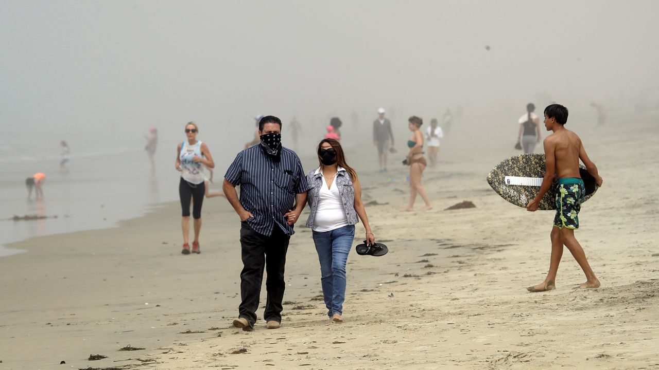 In this April 26, 2020 file photo people wear masks as they walk on the beach in Huntington Beach, Calif. Days after his predecessor abruptly quit, Orange County's new interim health officer will lift a requirement that residents wear face coverings in public and instead recommend they use masks to help prevent the spread of the coronavirus, an official said. (AP Photo/Marcio Jose Sanchez,File)