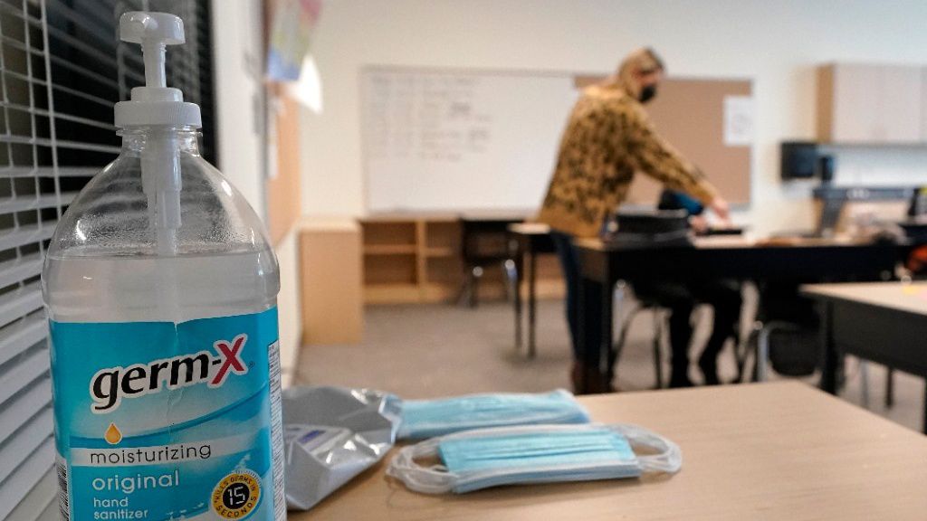 A tub of hand sanitizer sits on a desk with packs of masks, as children and teachers work at desks in the background.