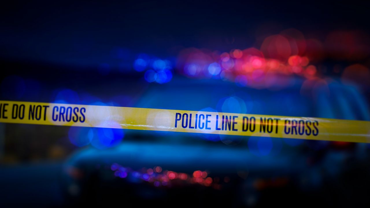 A stock photo of a Police Line "Do Not Cross" caution tape with a defocused police car with sirens flashing red and blue.