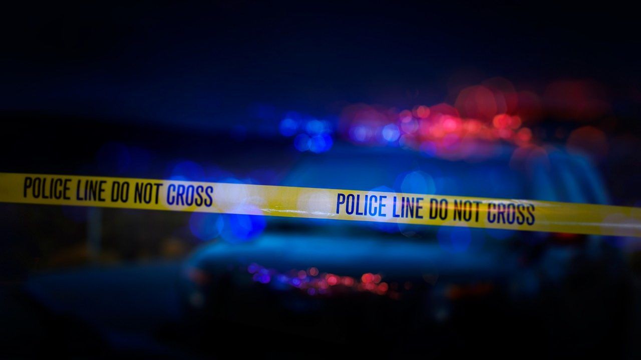 A stock photo of a Police Line "Do Not Cross" caution tape with a defocused police car with sirens flashing red and blue.