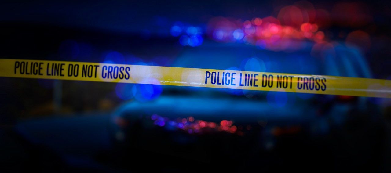 A stock photo of a Police Line "Do Not Cross" caution tape with a defocused police car with sirens flashing red and blue.