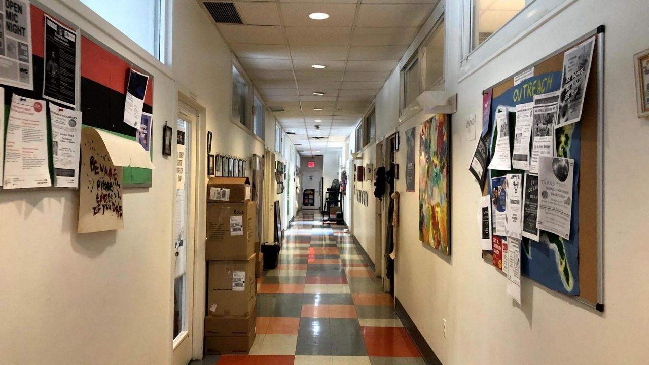 A hallway with multicolored floor tiled, bulletin boards with advertisements for events like Open Mic Night and doors to rooms for classes. 