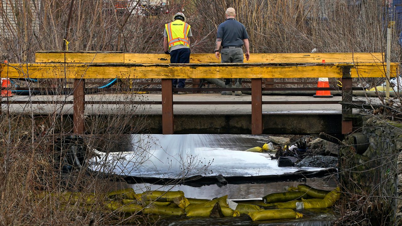 two men, one wearing a safety vest, stand on a bridge over a creek